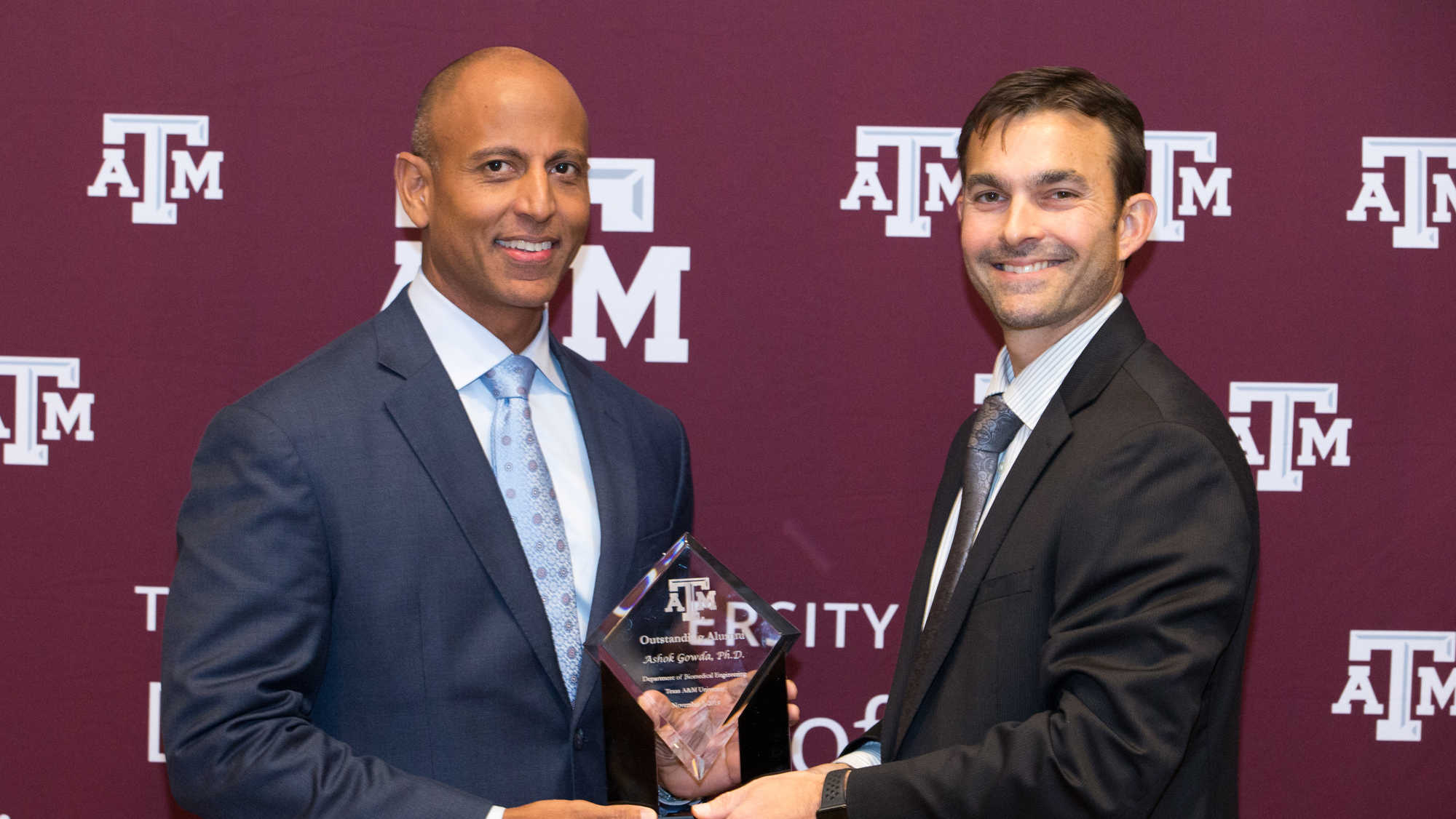 Two men in suits standing in front of maroon banner with white Department of Biomedical Engineering logo. They are holding a trophy made from clear crystal with the words 