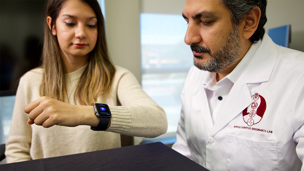 Student and professor in a laboratory both look at a smartwatch on the student's wrist.