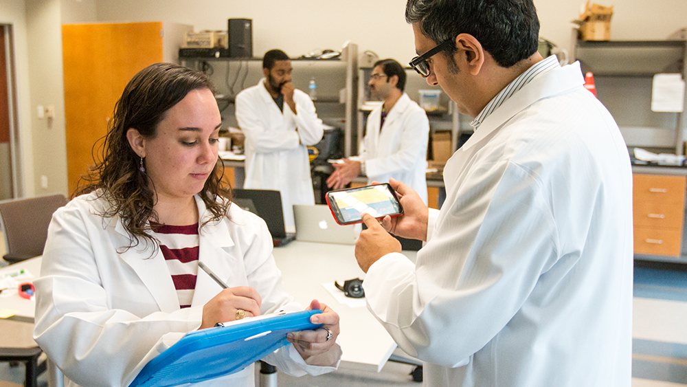 Dr. Sasangohar looks at a phone while a student holding a clipboard takes notes.