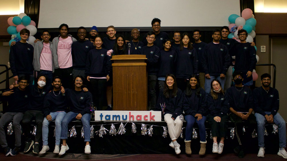 A group of students on a stage smiling at the camera. One half of the group are sitting on the edge of the stage and the other half are standing behind them. There is a podium in the middle of the stage with a sign that says 
