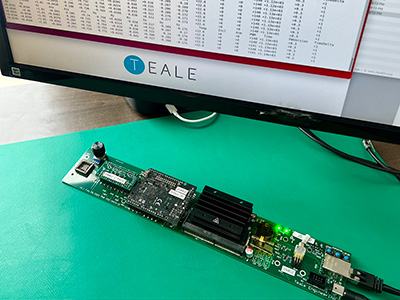 Hardware device with exposed circuitry lying on a desktop with a computer screen above it showing numerical readouts