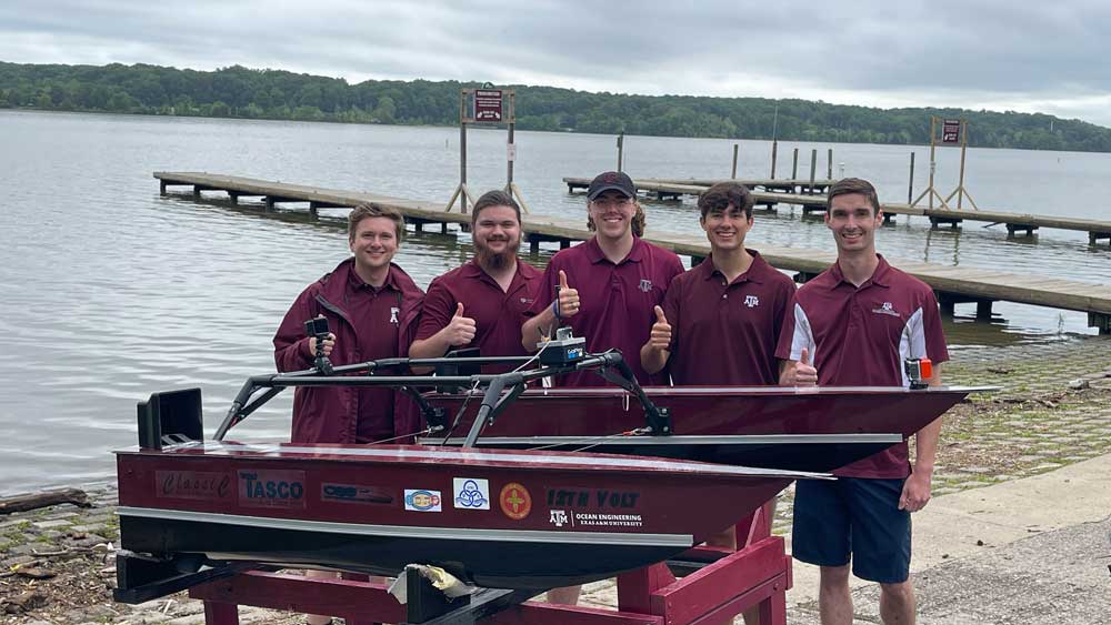 The five members of the Electric Boat Team stand behind their electric boat design and give thumbs up with Pohick Bay in the background.